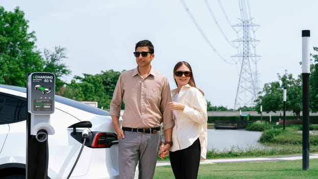 Young couple recharge EV car battery at charging station connected to power grid tower electrical industrial facility as electrical industry for eco friendly vehicle utilization. Expedient