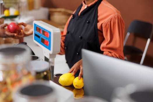 An African American woman in an apron assists a customer at a food market checkout counter. Fresh fruits and vegetables promote healthy eating. Technology aids in the buying process.