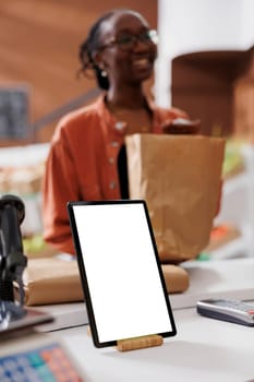 Closeup of digital tablet with blank white screen vertically placed on checkout counter at sustainable bio food market. Device displaying copy space mockup template while customer carries grocery bag.
