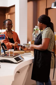 African American female client smiles while using her credit card for cashless payment. The local food market focuses on locally grown organic produce and eco friendly practices.
