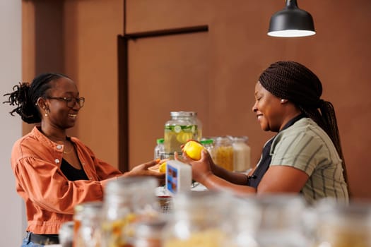 Cheerful african american woman assists a customer with freshly harvested produce at the grocery store cashier desk. Black vendor taking fresh lemons from client for weighing and packaging.