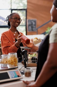 An African American woman sells freshly harvested organic produce to a customer in a grocery market. Female vendor receiving bottled sauce from smiling customer a checkout counter.
