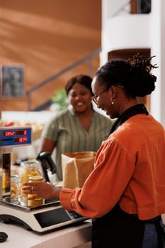 Female customer at a grocery store checkout counter with a smiling storekeeper weighing products on digital scale. Friendly vendor measuring weight of bio food items while client waits at desk.
