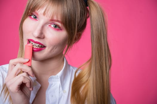 Portrait of a young woman with braces and bright makeup chewing gum on a pink background