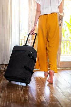 Unrecognizable woman walking out of hotel bedroom with suitcase. Vertical image. Vacation, trip concept.