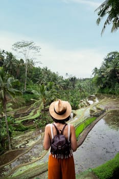 Back view of female tourist enjoying Tegalalang rice terrace landscape in Bali, Indonesia. Woman enjoying vacation and freedom in nature. Copy space. Vertical. Trip, vacation, mental health and healthy lifestyle concept.