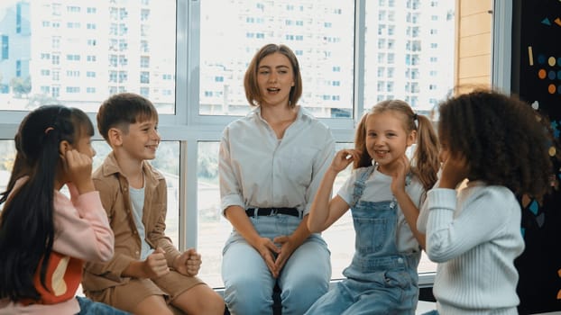 Smart caucasian teacher teaching while sitting on floor together surrounded by diverse students. Professional instructor playing with multicultural learner. Creative education concept. Erudition.