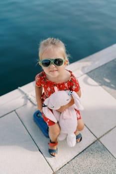 Little girl with a pink toy rabbit sits on a bollard on the pier and looks up. Top view. High quality photo