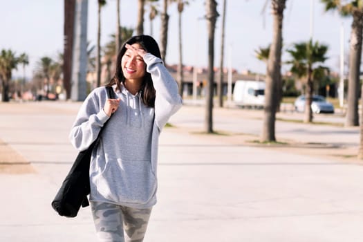 smiling young asian woman dressed in casual clothes walking with yoga mat hanging on her arm, sport and healthy lifestyle concept, copy space for text