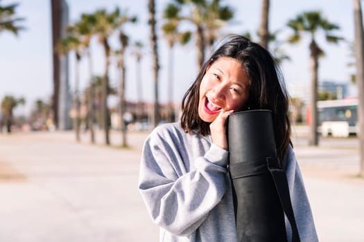 portrait of a young asian woman dressed in casual clothes laughing looking at camera while holding her yoga mat, healthy and active lifestyle concept, copy space for text