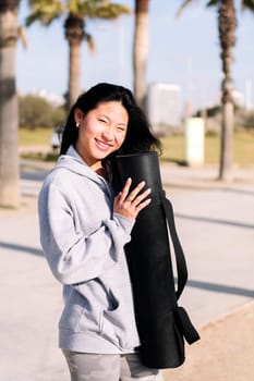 portrait of a young asian woman dressed in casual clothes smiling looking at camera while holding her yoga mat, healthy and active lifestyle concept, copy space for text