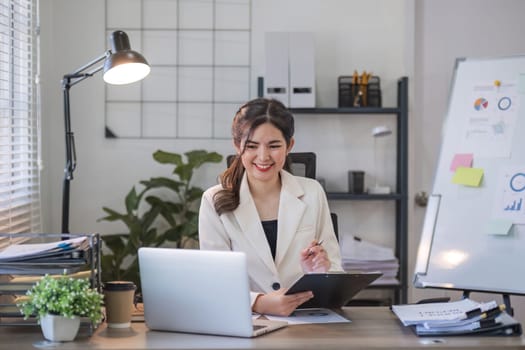 Asian businesswoman or accountant happily sitting and working with laptop on finance and business administration. On the desk in the office decorated with green plants..