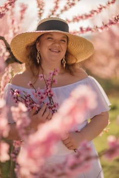 Woman blooming peach orchard. Against the backdrop of a picturesque peach orchard, a woman in a long white dress and hat enjoys a peaceful walk in the park, surrounded by the beauty of nature