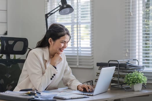 Asian businesswoman or accountant happily sitting and working with laptop on finance and business administration. On the desk in the office decorated with green plants..