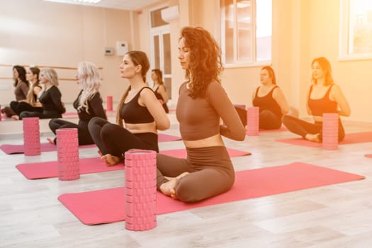 A group of six athletic women doing pilates or yoga on pink mats in front of a window in a beige loft studio interior. Teamwork, good mood and healthy lifestyle concept