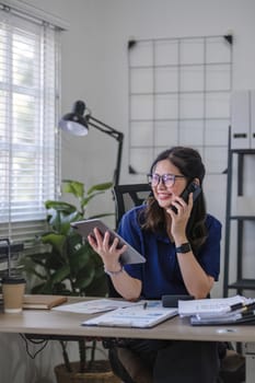 Young Asian business woman sits on the phone in an online business meeting using a laptop in a modern home office decorated with shady green plants..