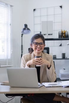 Asian businesswoman or accountant happily sitting and working with laptop on finance and business administration. On the desk in the office decorated with green plants..