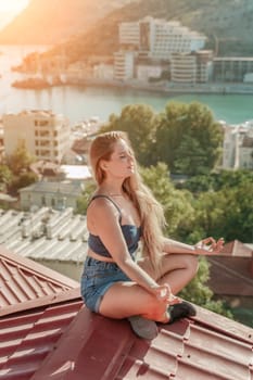 Woman sits on rooftop, enjoys town view and sea mountains. Peaceful rooftop relaxation. Below her, there is a town with several boats visible in the water. Rooftop vantage point