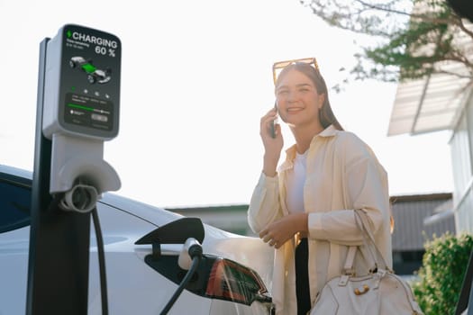 Young woman holding shopping bag talking on the phone while recharging EV car battery from charging station at city mall parking lot. Modern woman go shopping by eco car. Expedient