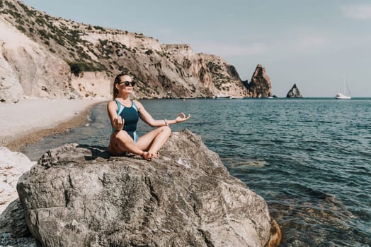 Yoga on the beach. A happy woman meditating in a yoga pose on the beach, surrounded by the ocean and rock mountains, promoting a healthy lifestyle outdoors in nature, and inspiring fitness concept