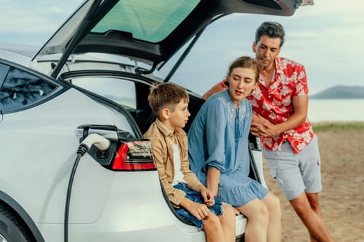 Family vacation trip traveling by the beach with electric car, lovely family sit on the trunk, charging EV car battery with green and sustainable energy. Family travel and eco-friendly car. Perpetual