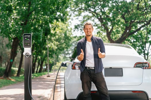 Man recharging battery for electric car during road trip travel EV car in natural forest or national park. Eco friendly travel during vacation and holiday. Exalt