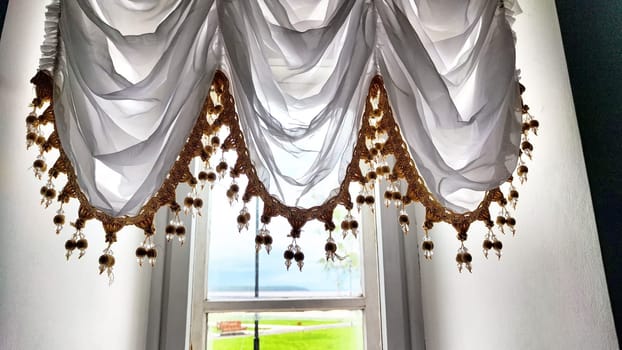 Elegant Window Drapes With Tassel Trim Overlooking a Sunny Day. White sheer curtains with decorative tassels framing a clear view of a blue sky. Background, texture, pattern, frame