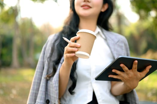 Satisfied young businesswoman drinking coffee and using digital tablet in the park.