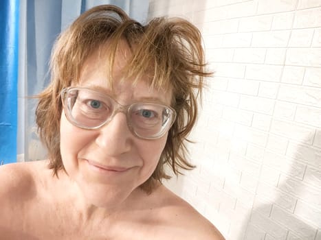 Shaggy cheerful middle aged woman with glasses smiling in bathroom in early morning. Smiling Woman Relaxing and taking selfie in mirror