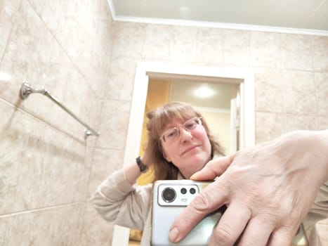 Mature woman captures her reflection while taking selfie. The reflection of middle aged girl with a phone in the mirror. Partial focus