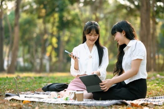 Two smiling female coworkers using digital tablet on grass in the city park.