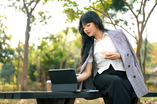 Young stylish woman eating sandwich and using digital tablet on park bench.