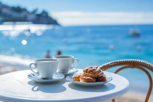Two azure coffee cups and a plate of pastries sit on a table overlooking the ocean, under a clear blue sky