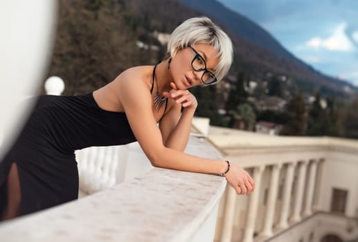 Portrait of an adult beautiful woman in glasses and an evening open dress on the street against the background of an ancient hotel