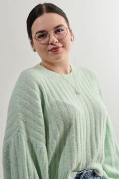 Portrait of beautiful female pupil in oversized sweater, wears spectacles, has pleased expression, has spare time after classes, going to make home assigment, poses indoor against white wall