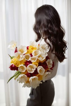 Beautiful curly woman with spring flowers tulips in hands looking at the window. Women's Day