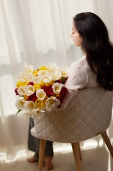 Beautiful woman with spring flowers tulips in hands sitting on chair near window. Women's Day