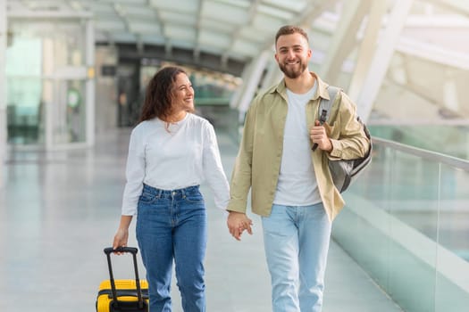 Portrait of romantic young couple walking together in airport terminal,, smiling millennial man and woman carrying luggage, holding hands and looking at camera, ready for honeymoon travel