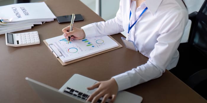 Finance concept. Close up businesswoman hold a graph pen and writing report, and memo, and analyzing business documents with a laptop computer.