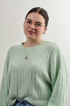 Portrait of beautiful female pupil in oversized sweater, wears spectacles, has pleased expression, has spare time after classes, going to make home assigment, poses indoor against white wall