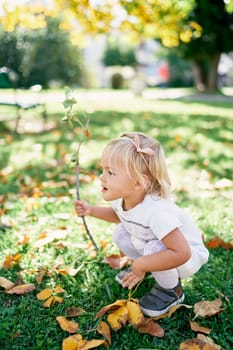 Little girl is squatting on a green lawn with a tree branch in her hand. High quality photo