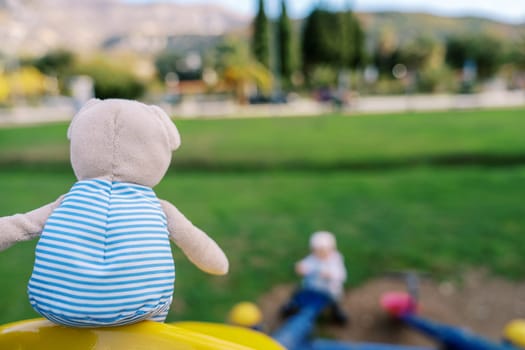 Teddy bear in a striped t-shirt sits on a swing in the playground. Back view. High quality photo
