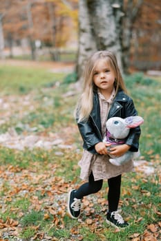 Little girl with a gray toy rabbit in her hands stands in the autumn forest. High quality photo
