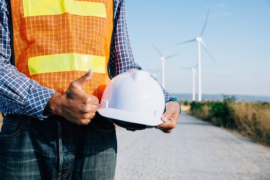 Engineer stands at windmill field holding safety helmet. Symbolizes renewable success innovation addressing global warming. Demonstrates industry leadership safety commitment.