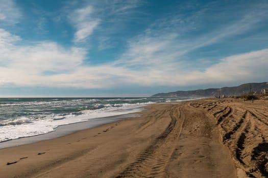 Empty beach scene with waves, tire tracks on sand, stretching to distant mountains under blue sky.