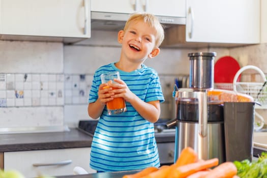 Smiling boy holding glass of carrot juice in kitchen. Healthy kids nutrition and homemade juice concept. Child making juice with juicer. Design for poster, banner, flyer