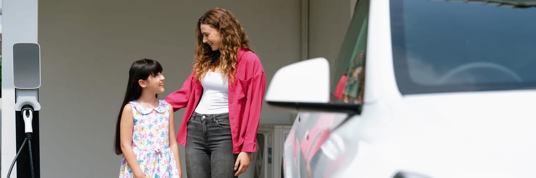 Happy little young girl learn about eco-friendly and energy sustainability as she help her mother recharge electric vehicle from home EV charging station. EV car and modern family. Panorama Synchronos