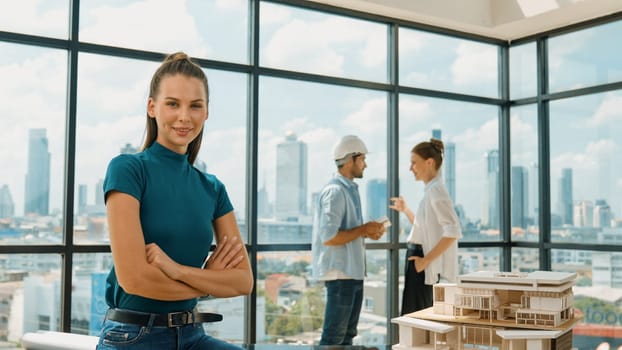 Young beautiful interior designer crossing arms while engineer team talking about house design. Skilled businesswoman smiling at camera with confident while sitting near house model. Tracery