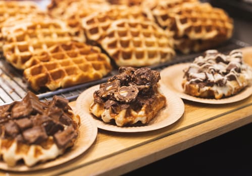 Appetizing fresh Belgian waffles with pieces of chocolate in cardboard eco-friendly plates lie on a wooden bakery display case, close-up side view with depth of field.