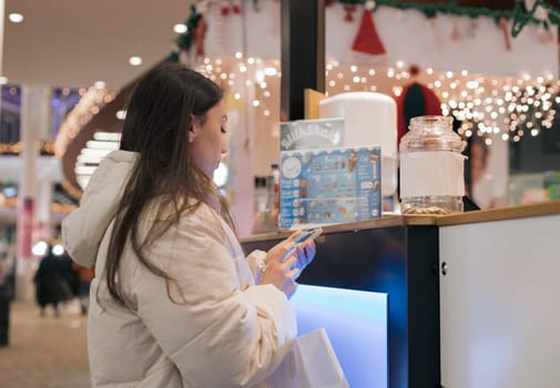 One beautiful young caucasian brunette girl in a white down jacket stands half-turned and looks into a smartphone behind a counter in a shopping center buying sweets, close-up side view. Offline shopping concept.